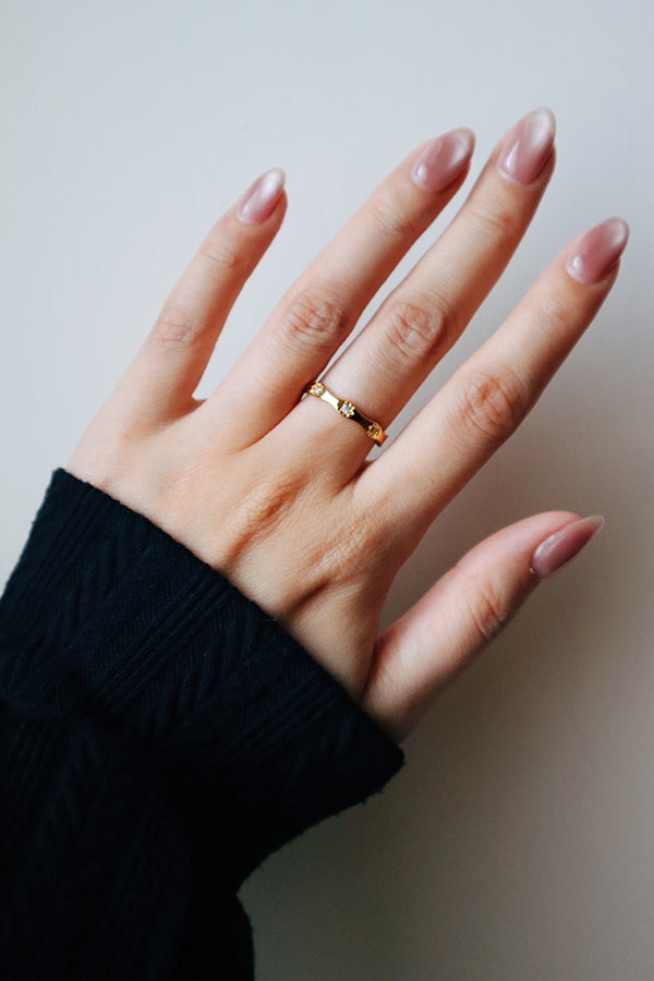 A close-up of a woman's hand with long, nude-toned manicured nails, showcasing the Amara Sparkle Ring on her ring finger. This gold band features a slightly wavy design embedded with three small, sparkling stones. She is wearing a black long-sleeved top.