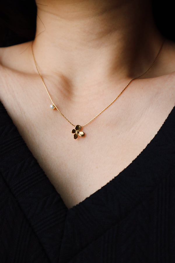 A close-up of a woman's collarbone and chest, showcasing the Chloe Bloom Pendant Necklace. This delicate gold chain holds a small, sculpted four-petal flower pendant and a subtle single pearl accent near the collarbone. The woman is wearing a black V-neck top.
