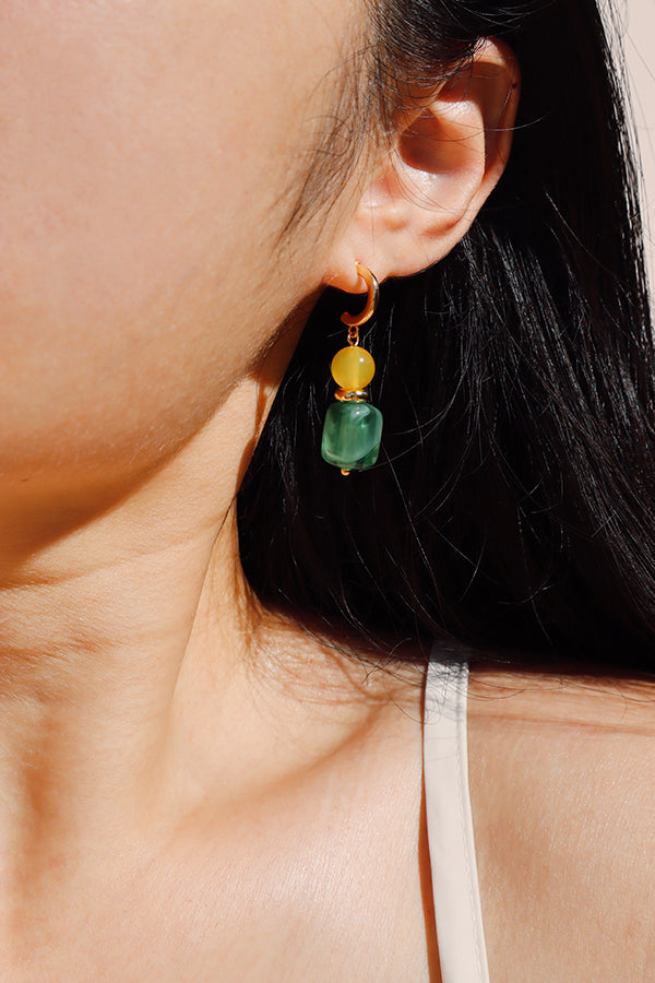 A close-up side profile of a woman's ear and neck, showcasing a gold hoop drop earring with a bright yellow round bead and a marbled green rectangular bead. Sunlight casts a pronounced shadow below the jawline, highlighting the earring's vibrant colors.