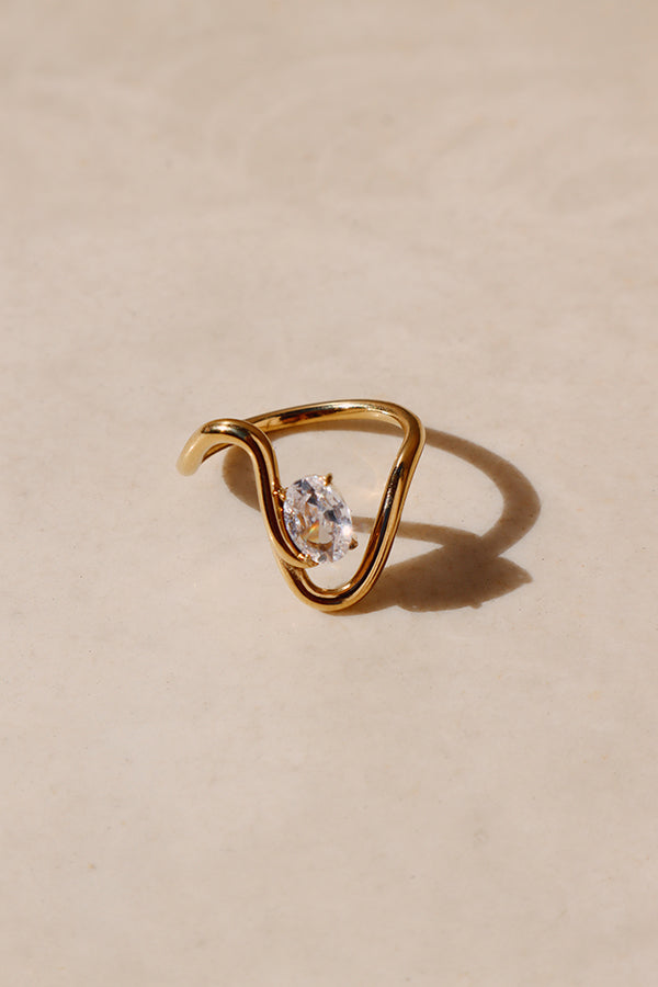 An overhead, high-contrast shot of the Eveline Curve Ring, a bold gold statement ring featuring a swirling, asymmetric band that cradles a sparkling oval cubic zirconia stone. The ring is casting a sharp shadow on a light beige surface.