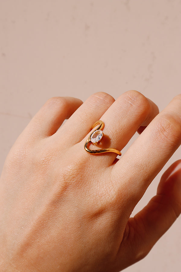 A close-up of a woman's hand, showcasing the Eveline Curve Ring worn on her ring finger. The gold ring features a curved, wavy band cradling a brilliant clear stone. The background is a soft, light pink.