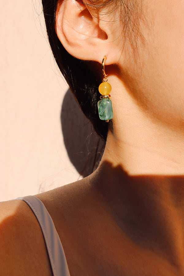 A close-up side profile of a woman's ear and neck, featuring the Gia Sunlit Oasis Earring. This beaded drop earring features bright yellow and marbled green stones hanging from a gold hoop, catching the bright sunlight.