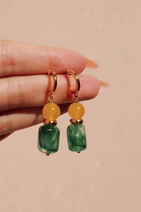 A female hand with light-colored, long nails holds a pair of Gia Sunlit Oasis Earrings. These colorful drop hoops feature a bright yellow round bead and a rectangular green marbled resin bead, set against a soft, pink background.