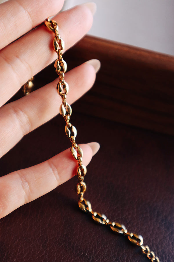 A female hand with long, almond-shaped nails delicately holds a segment of the gold-toned coffee-bean link bracelet, emphasizing the shine and structure of the links against a dark, warm background.