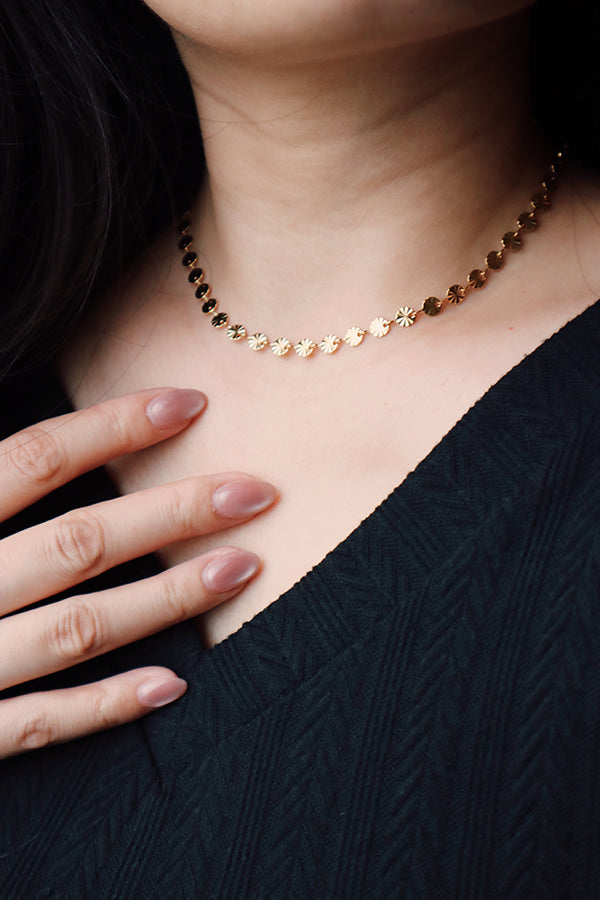A close-up of a woman's neck and chest, featuring a delicate gold flower-link choker. The woman's hand, with long, mauve-colored manicured nails, rests on her black, textured top, highlighting the gold choker.