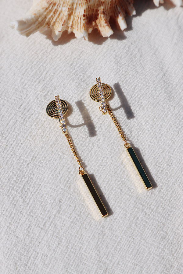 An overhead shot of a pair of gold bar drop earrings, each featuring a spiral-shaped top with pave-set stones and a delicate chain suspending a thin gold bar. They are resting on a white textured fabric, with a large seashell in the background. Sunlight casts sharp shadows.
