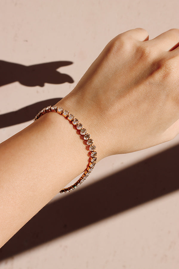 A close-up of a woman's wrist and arm, wearing a gold tennis bracelet adorned with a continuous line of clear, shimmering stones. The piece is highlighted by harsh, diagonal sunlight that casts a prominent shadow across the wrist.