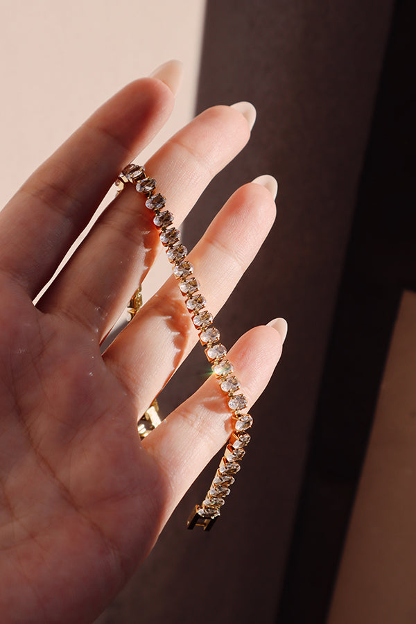 A female hand with long, light-colored manicured nails delicately holds the Harper Radiance Bracelet, allowing the light to catch the continuous line of sparkling white stones. The bracelet's gold clasp is visible near the fingers.