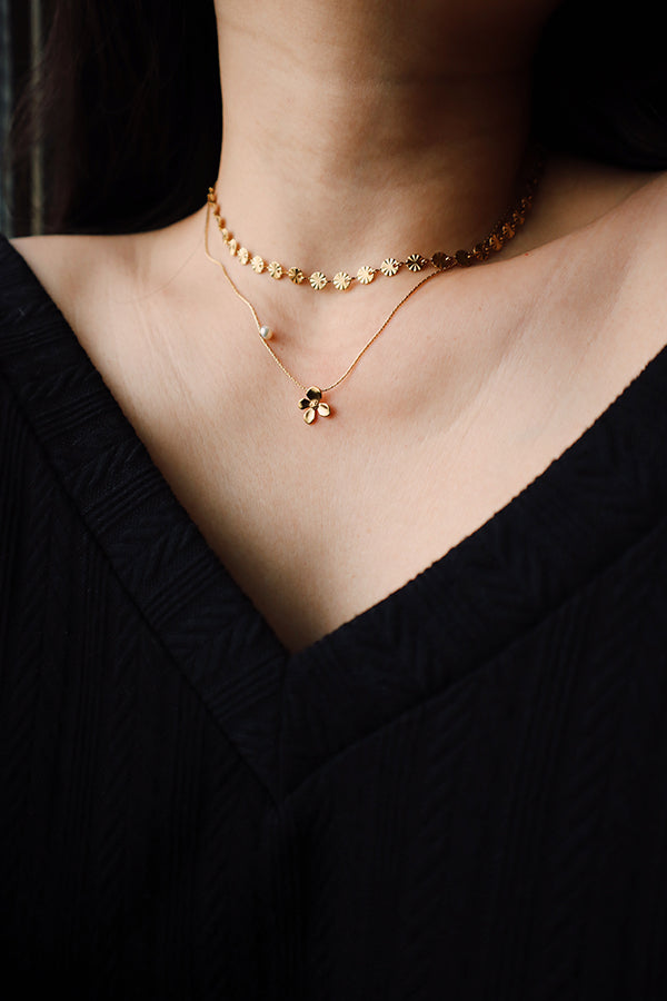 A close-up of a woman's neck and chest, featuring two layered gold necklaces: the Chloe Bloom Pendant Necklace and a shorter Sienna Glow Choker made of flower-shaped links. The layered look is set against a black V-neck top.