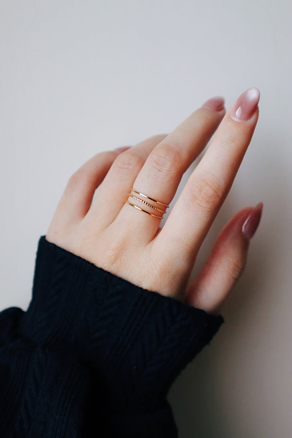 A woman's hand with long, pink-toned, iridescent manicured nails, prominently wearing the Liora Triple Band Ring on her middle finger. The ring features three separate gold bands designed for easy stacking. She is wearing a black long-sleeved top.