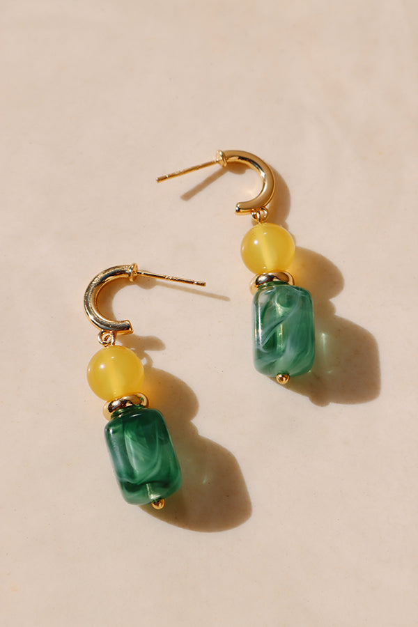 An overhead shot of a pair of Gia Sunlit Oasis Earrings with open gold hoops. Each earring features a yellow round bead and a marbled green rectangular bead. The earrings are resting on a warm, light-colored surface with soft shadows.