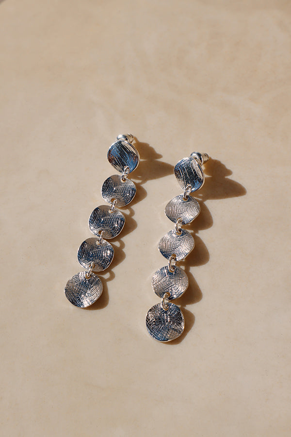An overhead shot of a pair of long, silver-toned drop earrings, each featuring five stacked, interconnected discs with a distinctive hammered or textured surface. The earrings are resting on a smooth, beige surface.
