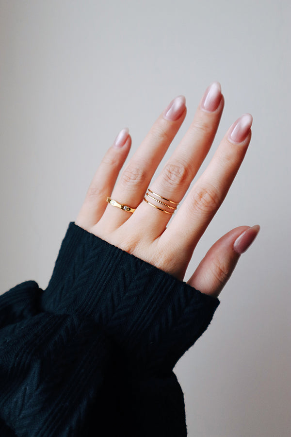 A woman's hand showing a ring stack across two fingers. The middle finger wears the Liora Triple Band Ring, while the index finger wears a simple gold band with small embedded stones (the Amara Sparkle Ring). The woman has long, iridescent manicured nails.