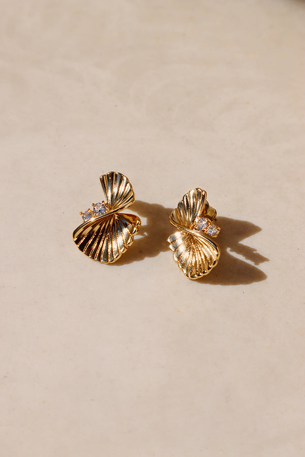 An overhead shot of a pair of Serena Wave Earrings. These gold stud earrings feature a fan-like, textured wave design, each embellished with three small, sparkling crystals. They are placed on a light, smooth surface, catching the light and casting soft shadows.