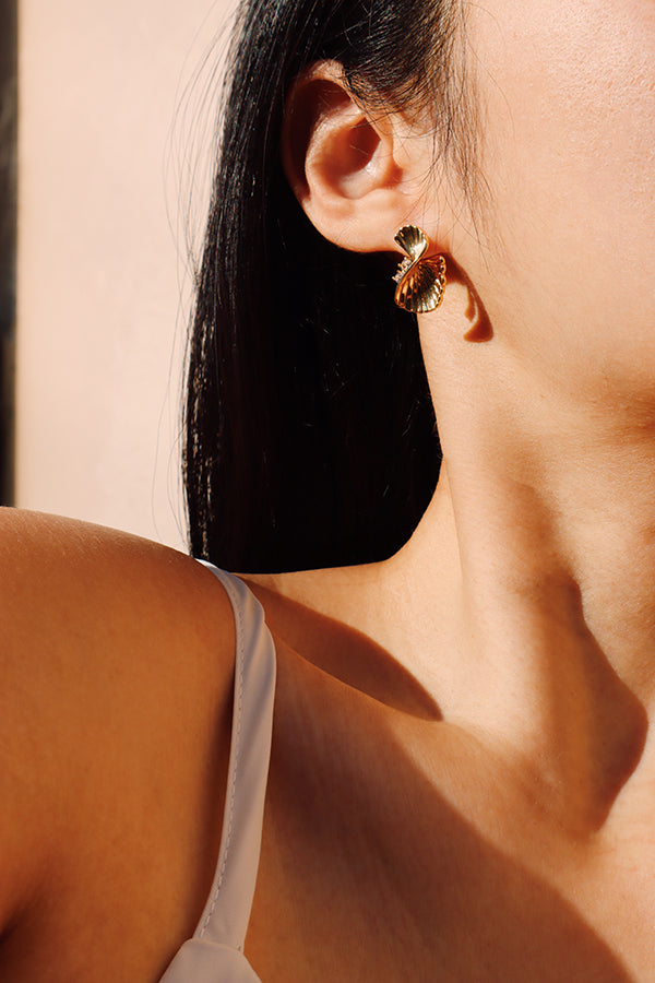 A close-up side profile of a woman's ear and neck, highlighting a single Serena Wave Earring. This gold stud earring features a fan-like, textured wave design that mimics a seashell, catching natural sunlight which casts shadows. The woman has dark hair and is wearing a white strap top.