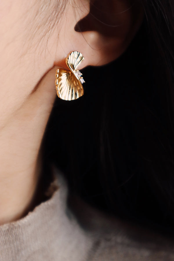 A close-up side profile of a woman's ear, highlighting a single Serena Wave Earring. This gold stud earring presents a fan-like, textured wave design with crystal accents, set against a dark, blurred background that emphasizes the earring's shine. The woman has dark hair and is wearing a neutral-toned top.