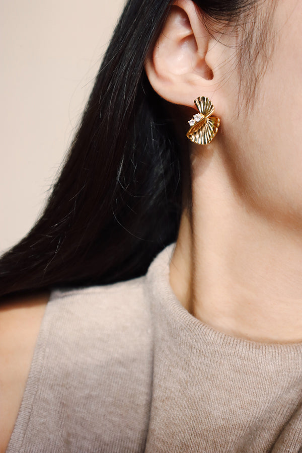 A close-up side profile of a woman's ear and neck, featuring a Serena Wave Earring. This gold stud earring has a fan-like, textured wave design and small crystal embellishments. The woman has long, dark hair pulled back slightly and is wearing a neutral-toned, ribbed top.