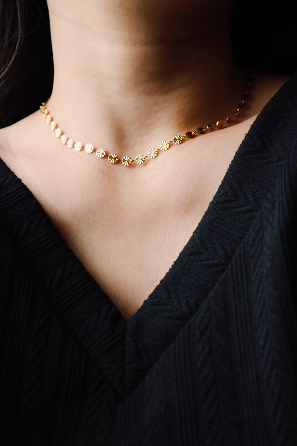 A close-up of a woman's neck and chest, showcasing the Sienna Glow Choker, a delicate gold chain necklace adorned with small, interconnected flower-shaped links. The choker rests above the V-neckline of a black, textured top.