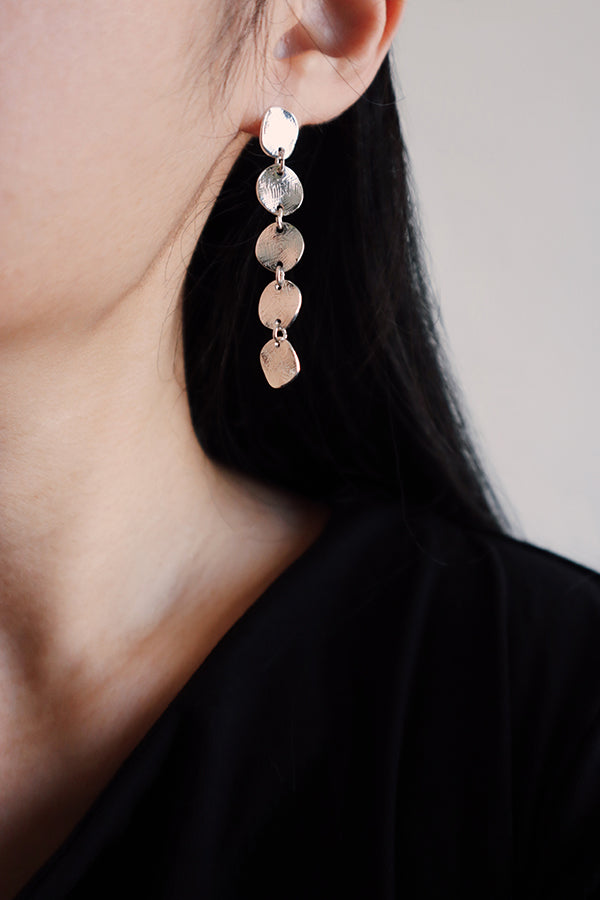 A close-up side profile of a woman's ear and neck, showcasing a long, dangling silver-toned earring made of five stacked, textured coin discs. The woman has long, dark hair and is wearing a black top.