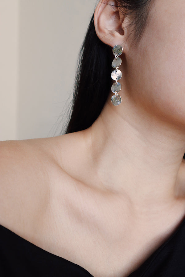 A close-up of a woman's ear, neck, and exposed shoulder, showcasing a long, dangling silver-toned earring made of five stacked, textured coin discs. The woman has long, dark hair and is wearing a black off-the-shoulder top.