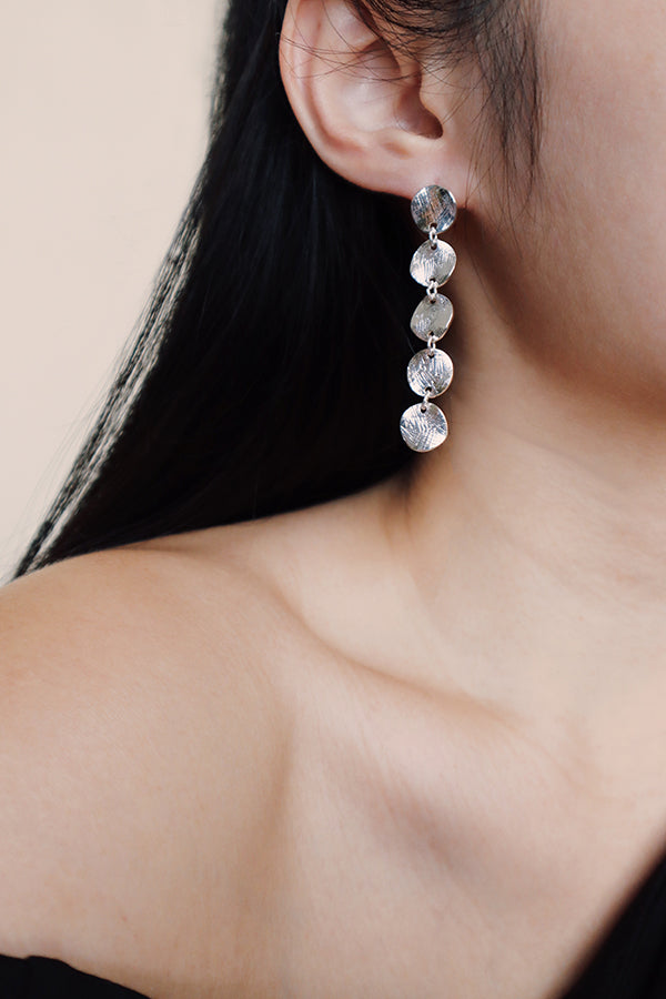 A close-up of a woman's ear, shoulder, and exposed collarbone, showcasing a long, dangling silver-toned earring made of five stacked, textured coin discs. The woman has long, dark hair.