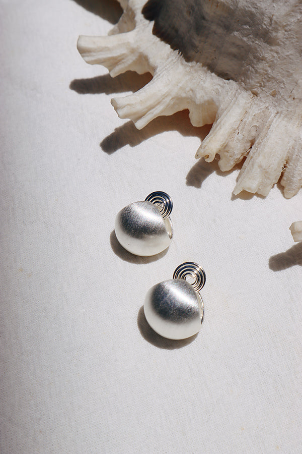 An overhead shot of a pair of silver-toned dome-shaped stud earrings with a brushed or textured finish, and a unique spiral backing visible. They are resting on a white textured fabric, with a large seashell partially visible in the top background.