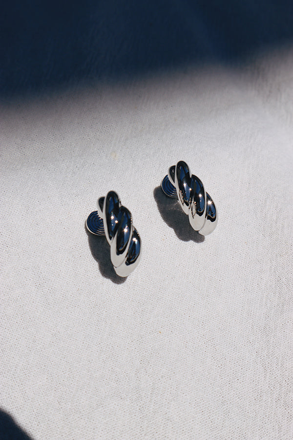 An overhead shot of a pair of shiny silver clip-on earrings, each featuring a twisted, croissant-like shape. They are resting on a white textured fabric, with sharp shadows cast by sunlight.