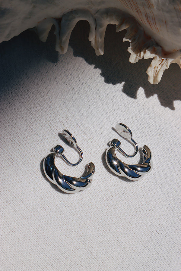 An overhead shot of a pair of shiny silver clip-on earrings, each featuring a twisted, croissant-like shape, with their clip mechanisms visible. They are resting on a white textured fabric, with a large seashell in the top background, and strong shadows cast by sunlight.