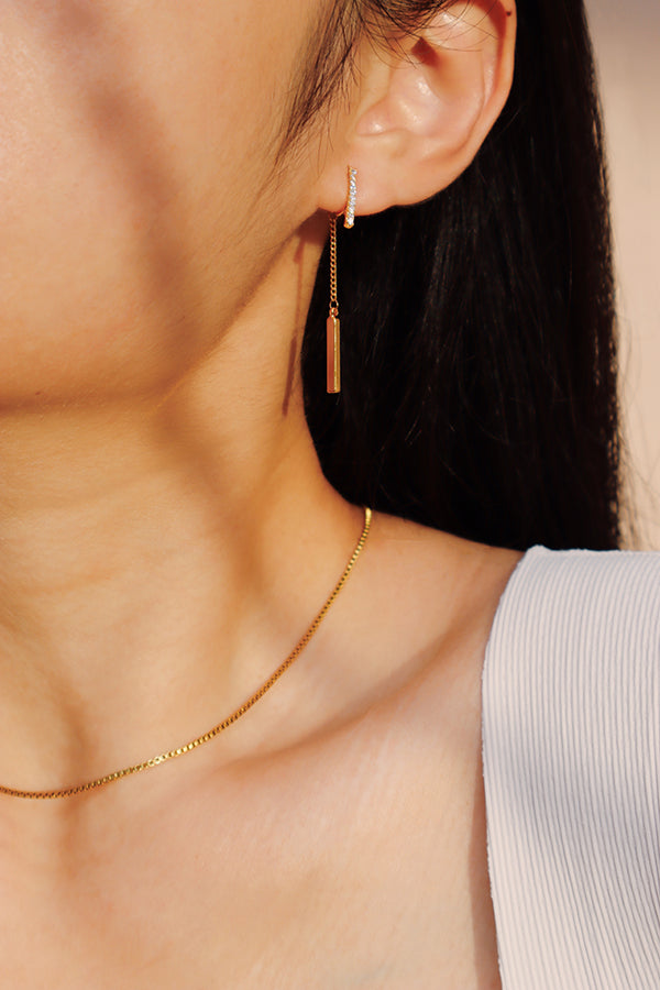 A close-up side profile of a woman's ear and shoulder, showcasing a gold hoop earring with pave-set stones, from which dangles a thin gold bar on a delicate chain. The woman has long dark hair and is wearing a white ribbed tank top. A thin gold chain necklace is also visible.
