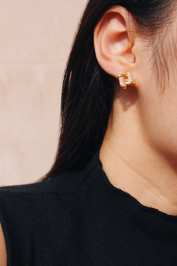 A close-up side profile of a woman's ear and shoulder, showcasing a small, gold-toned interlocking square stud earring, with one side covered in sparkling clear pave-set stones. The woman has long dark hair and is wearing a black ribbed turtleneck.