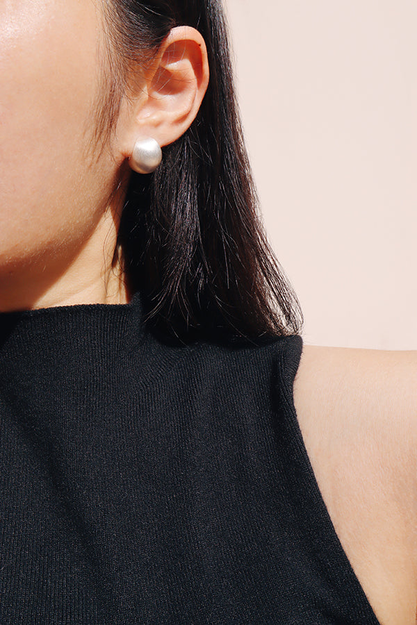 A close-up side profile of a woman's ear and shoulder, showcasing a large, shiny silver dome-shaped stud earring. The woman has long dark hair and is wearing a black ribbed sleeveless top.