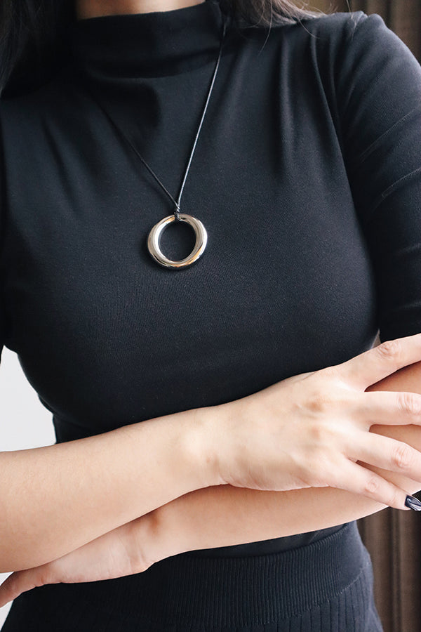 A close-up of a woman's chest and neck, wearing a black cord necklace with a large, hollow silver circular pendant. Her arms are crossed. The woman has dark hair and is wearing a black turtleneck top and a ribbed black skirt.