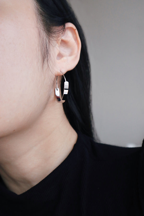 A close-up side profile of a woman's ear, showcasing a small, shiny silver hoop earring with a flattened, open design. The woman has long dark hair flowing around her ear and is wearing a black ribbed top.