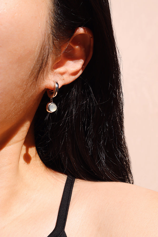 A close-up side profile of a woman's ear, showcasing a shiny silver small hoop earring from which a round, iridescent white opalite bead dangles. The woman has long dark hair and is wearing a black tank top.