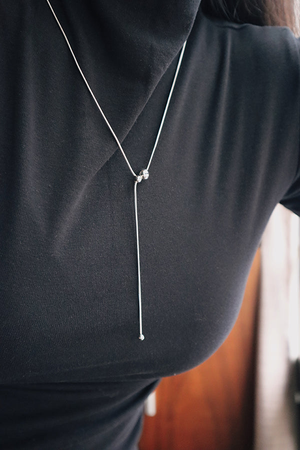 A close-up of a woman's chest and neck, wearing a delicate silver lariat necklace. The necklace features a sliding bead that adjusts the drop length, ending in a small bead. The woman is wearing a black turtleneck.