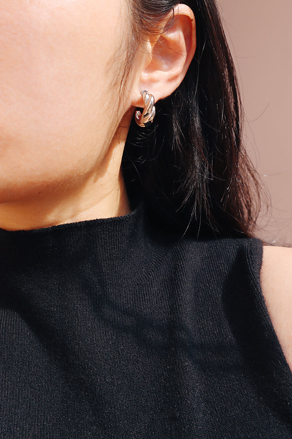 A close-up side profile of a woman's ear, showcasing a small, shiny silver twisted hoop or clip-on earring. The woman has long dark hair flowing around her ear and is wearing a black ribbed turtleneck.