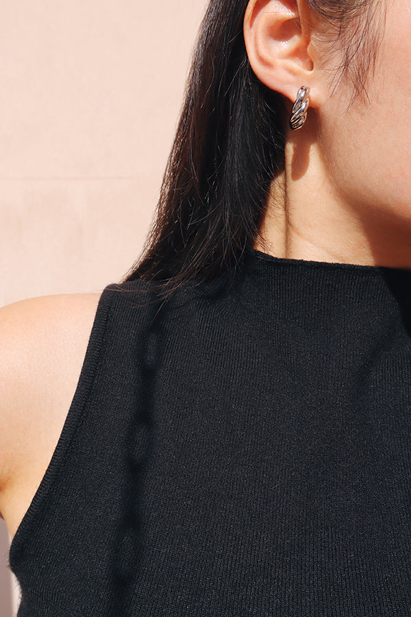 A close-up side profile of a woman's ear, showcasing a small, shiny silver twisted hoop or clip-on earring. The woman has long dark hair and is wearing a black ribbed sleeveless top.