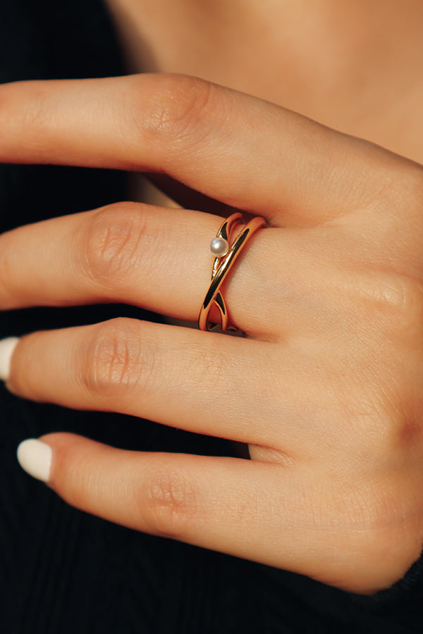 A woman's hand adorned with a delicate gold wrap ring featuring a single pearl, styled against a black outfit for an elegant touch.