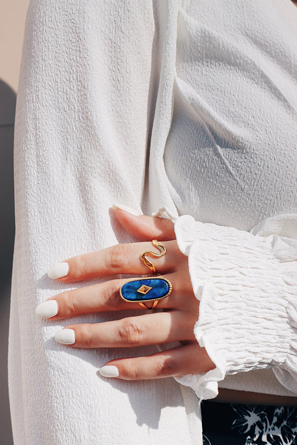 A woman in a white textured blouse wearing a bold lapis lazuli statement ring and a wavy gold ring, highlighting boho-chic jewelry styling.