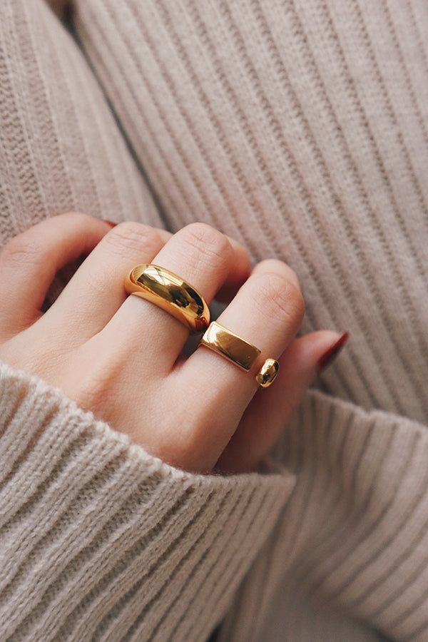 Close-up of a hand wearing two bold gold statement rings, resting on soft beige knit fabric, giving off a cozy and luxurious vibe.