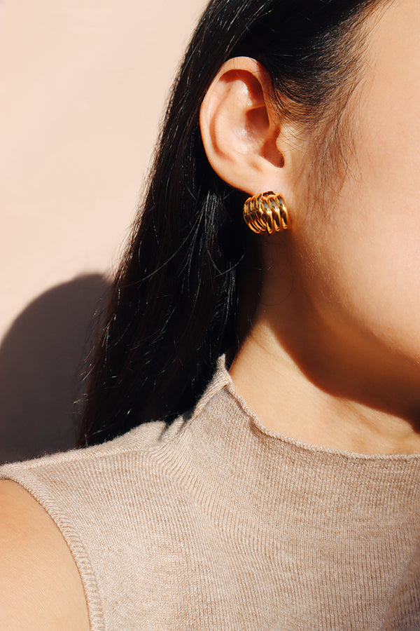 A woman's ear wearing vintage-inspired gold stud earrings with a modern, twisted design, against a beige background.