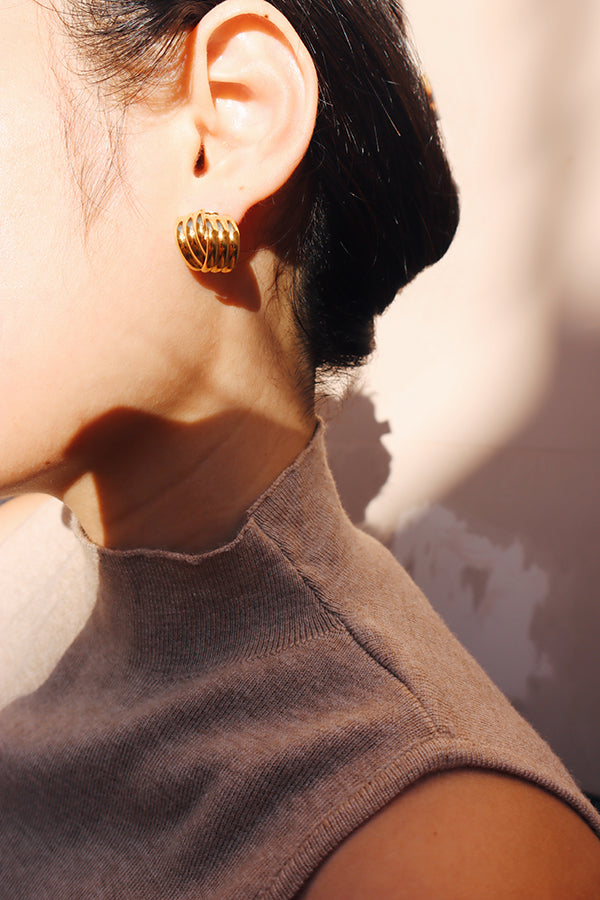 Close-up view of a gold vintage stud earring with a layered design, capturing sunlight against a beige sleeveless top, highlighting its warm shine and classic style.