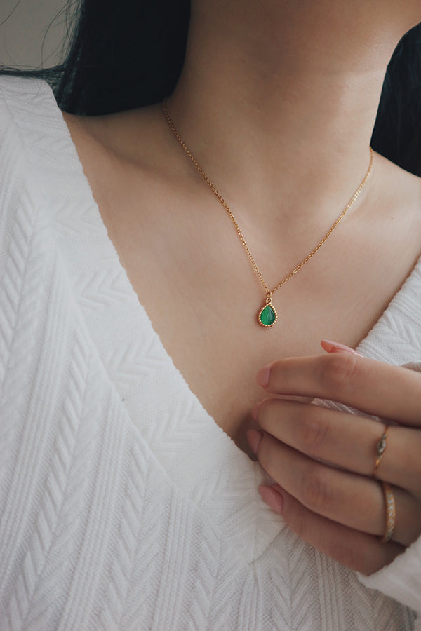 A close-up of a woman wearing a colorful gem necklace featuring a green cat's eye teardrop pendant, styled with a white textured top, ideal for spring outfits.