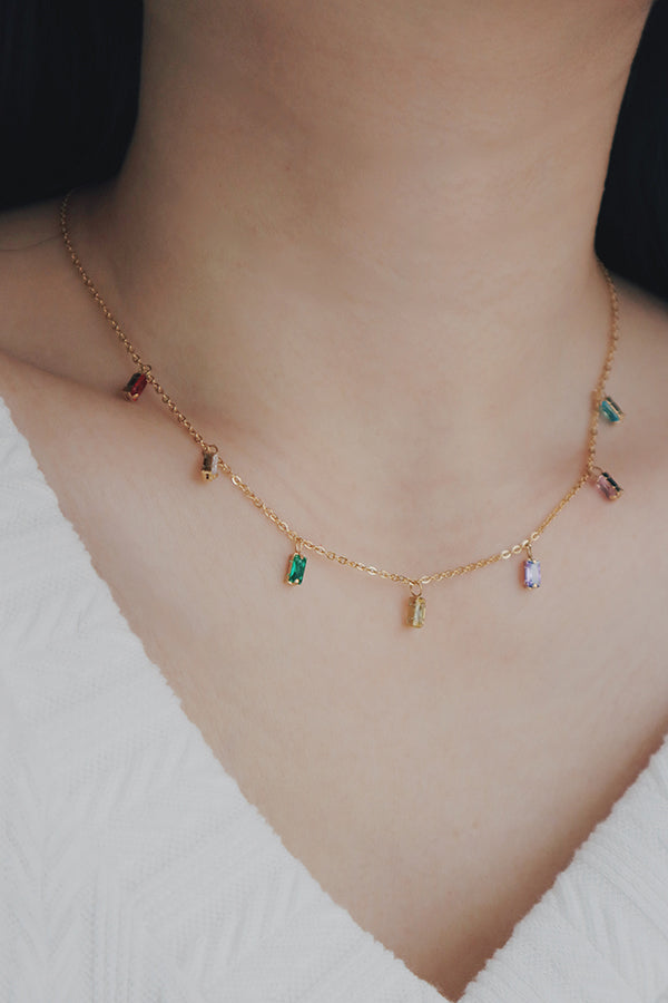 A close-up shot of a woman modeling a delicate gold chain necklace, featuring an array of rectangular cubic zirconia stones in bright, assorted colors such as red, blue, green, and purple. Each stone is individually set, creating a playful yet elegant piece suitable for both casual and formal wear.