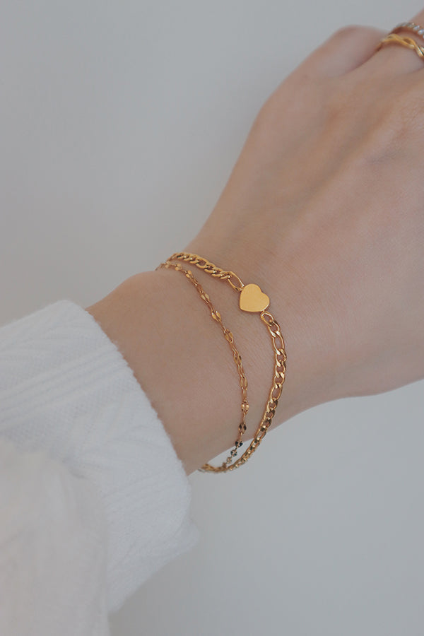 A close-up of a wrist adorned with a delicate gold layered bracelet featuring a small heart charm. The bracelet consists of two fine chains, one plain and the other with the heart charm, creating a sweet and romantic look. The bracelet sits elegantly on the wrist, complemented by a soft, white knit sleeve, making it a perfect accessory for a feminine and charming style.