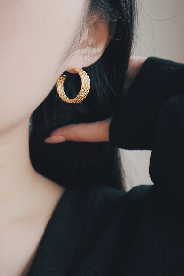 A detailed photo of textured gold-plated hoop earrings, displayed flat to showcase their intricate design. The earrings feature a raised, woven-like pattern that gives them a distinctive and fashionable look, suitable for both casual and formal outfits.