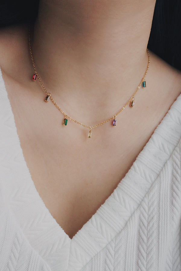 A close-up image of a womans neck wearing a dainty gold chain necklace adorned with colorful cubic zirconia stones in various shades, including red, green, yellow, and purple. The stones are evenly spaced, creating a subtle yet vibrant look, perfect for adding a pop of color to any outfit.