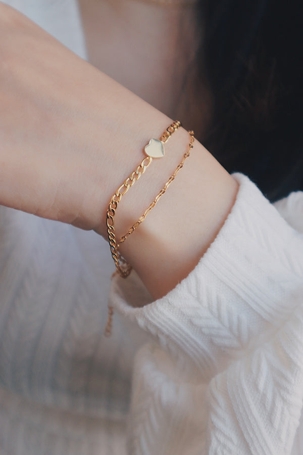 A wrist elegantly adorned with a gold layered bracelet featuring a heart charm. The bracelet's delicate chains and small heart detail add a touch of whimsy and romance, perfect for pairing with spring outfits. The light, neutral tones of the knit sleeve enhance the bracelet's soft and subtle aesthetic.